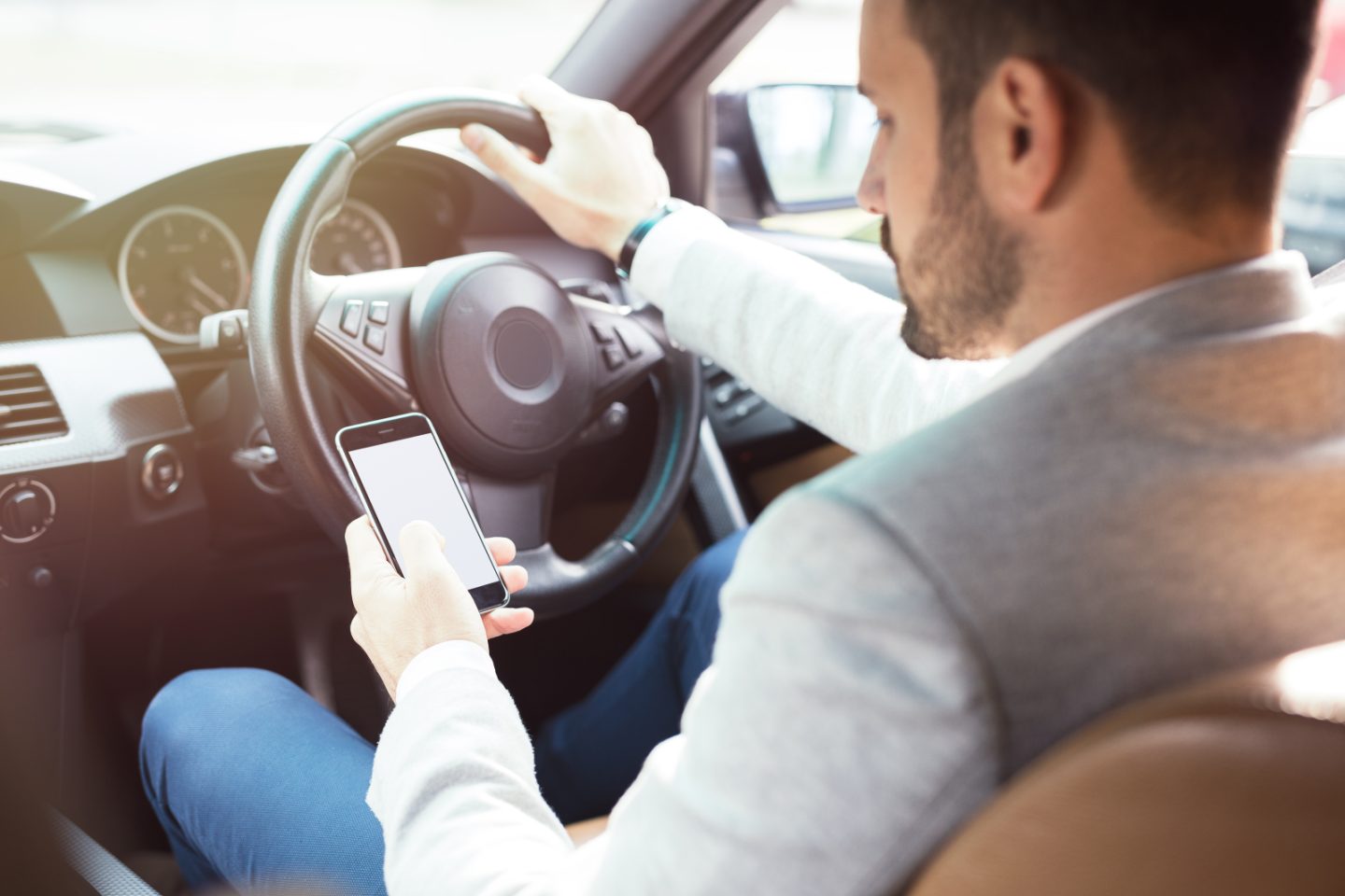 A man driving with one hand on the steering wheel. He has his mobile phone in his other hand and is looking at it.