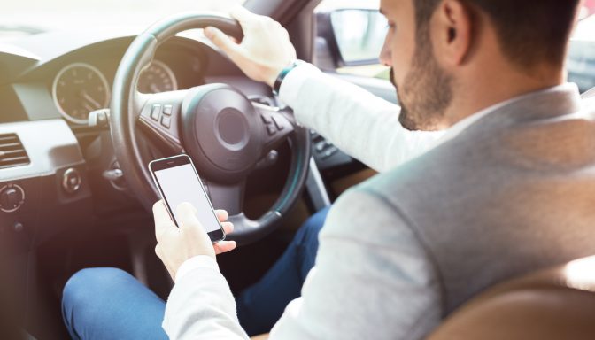 A man driving with one hand on the steering wheel. He has his mobile phone in his other hand and is looking at it.