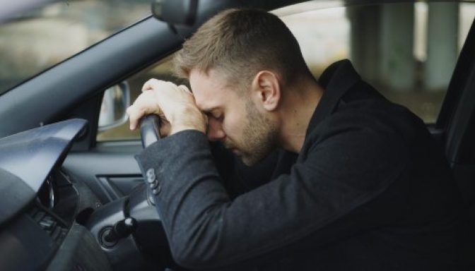 A man sat in his car, leaning onto the steering wheel, suggesting he is stressed or overwhelmed.