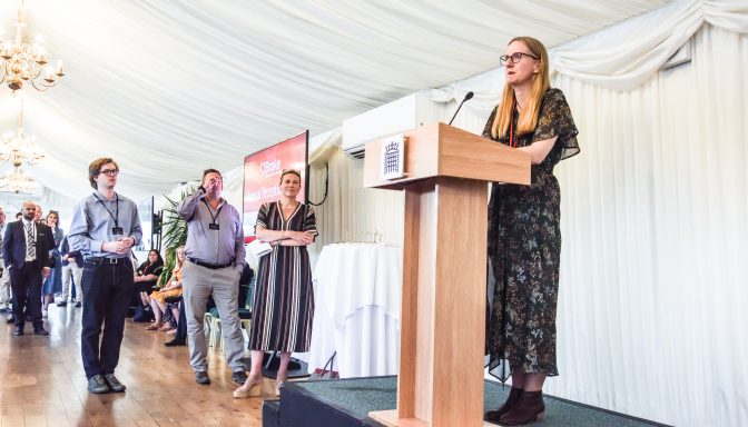 A middle aged woman in a dress is on stage, speaking at a lectern. People are stood around her listening.