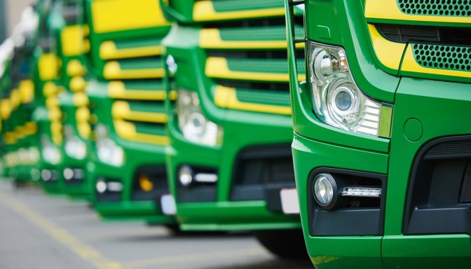 A fleet of green lorries parked in a row, showing their headlights and front bumpers.