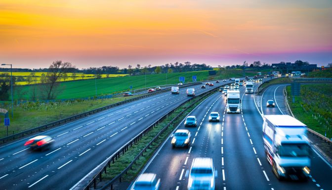 A busy four-lane motorway with fields either side. The sun is setting and vehicles have their headlights on.