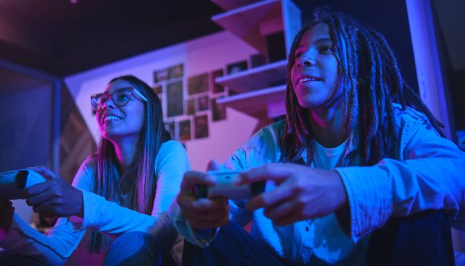 Two smiling young people sit next to each other on a sofa, playing a video game.