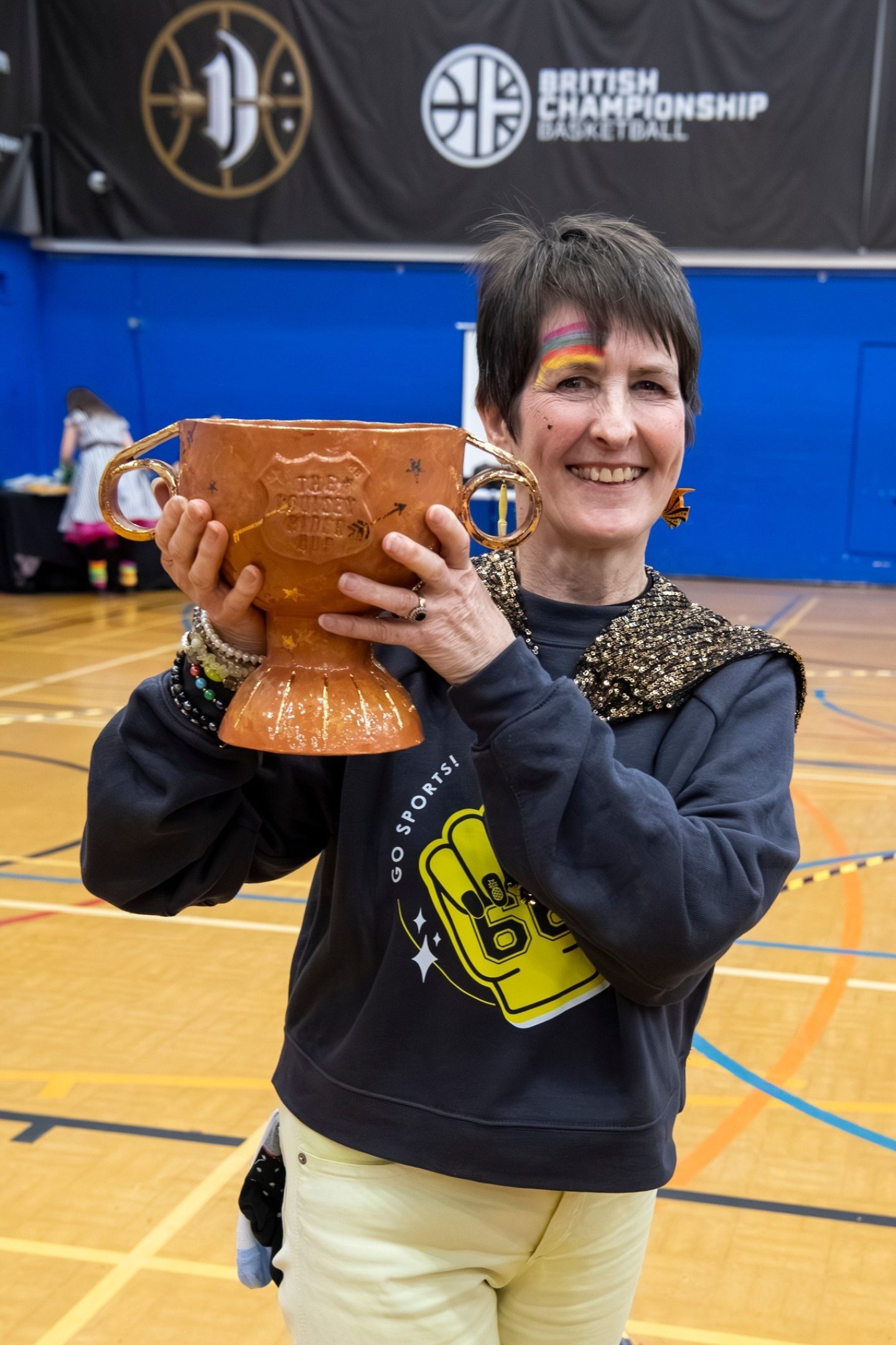 A middle aged woman wearing a black sweatshirt poses for a photo holding a ceramic trophy, embossed with the words 'The Louisey Rider Cup'.