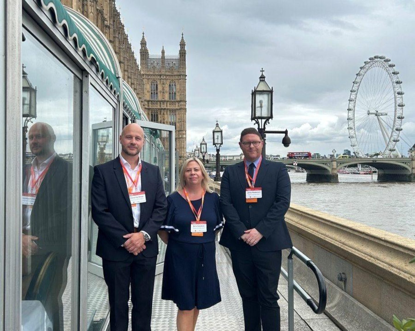 Three smartly dressed people pose for a photo on a balcony in Westminster. The London Eye can be seen in the background.