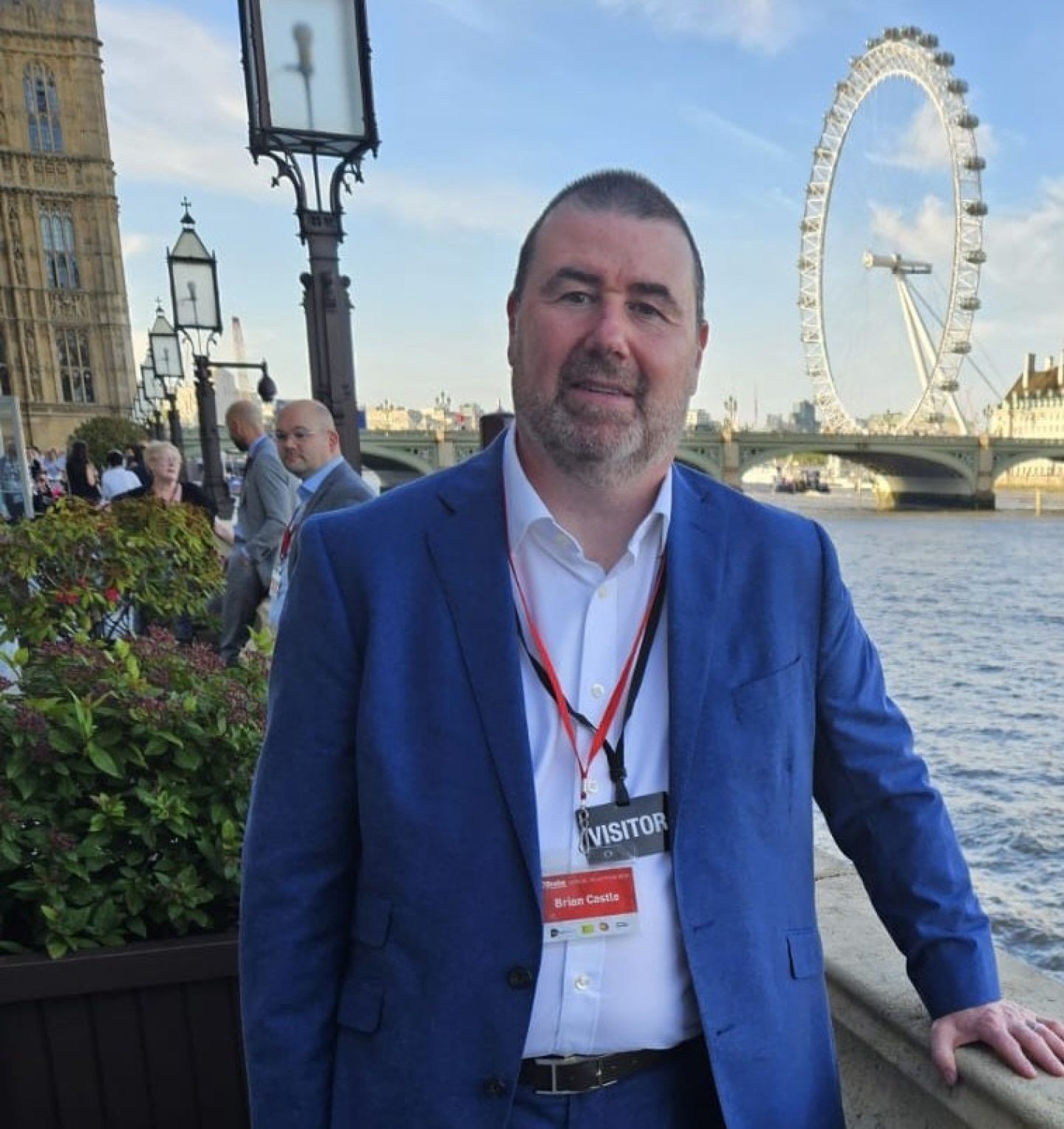 A man wearing a suit poses on a balcony by the River Thames. The London Eye can be seen in the background.