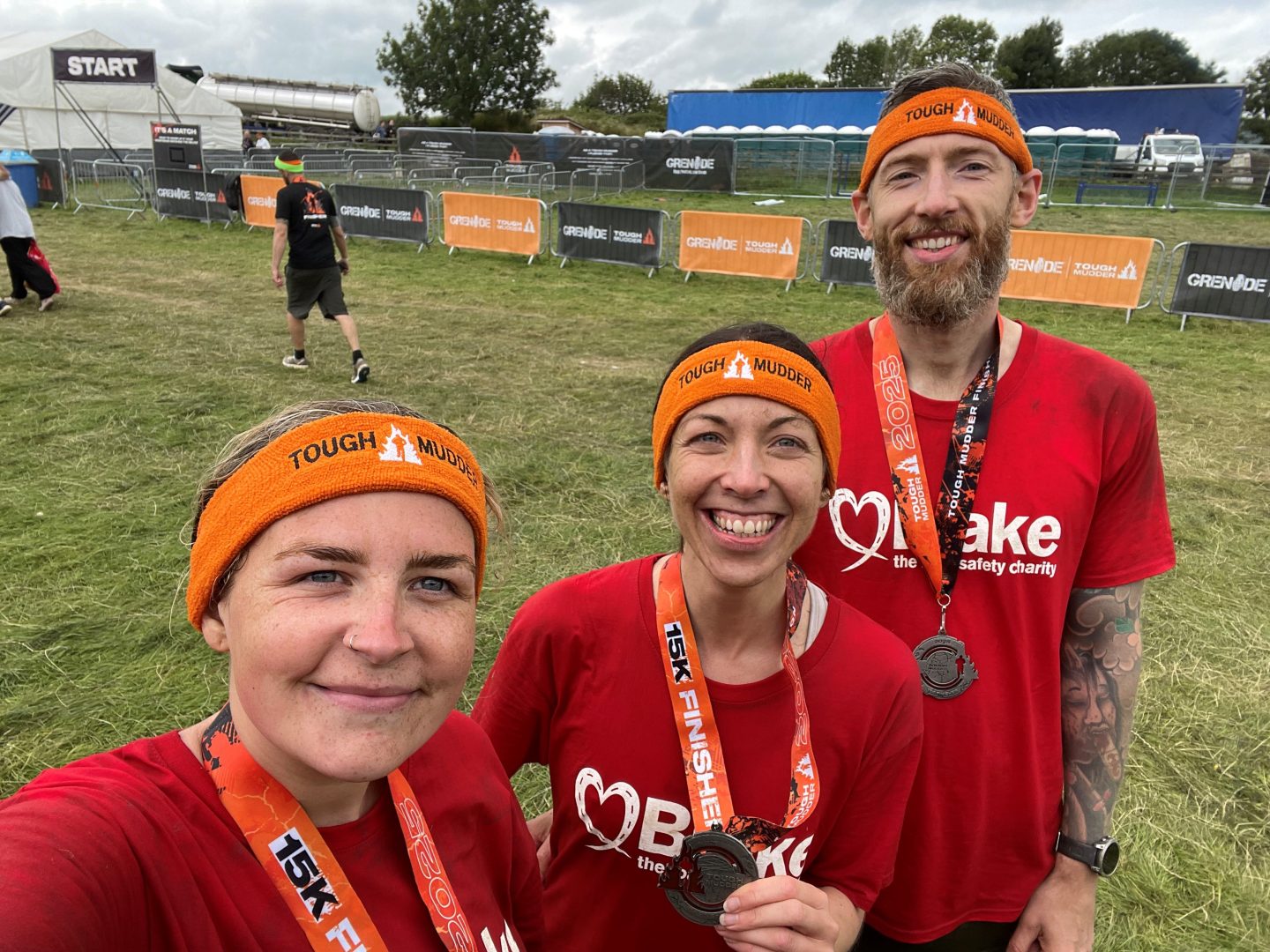 Three people pose for a selfie wearing t-shirts with Brake, the road safety charity's logo on. They have orange headbands and medals for a Tough Mudder event.