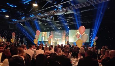 A large hall filled with people and illuminated by blue lights. A large screen shows the text "UK Fleet Champions Awards 2023"