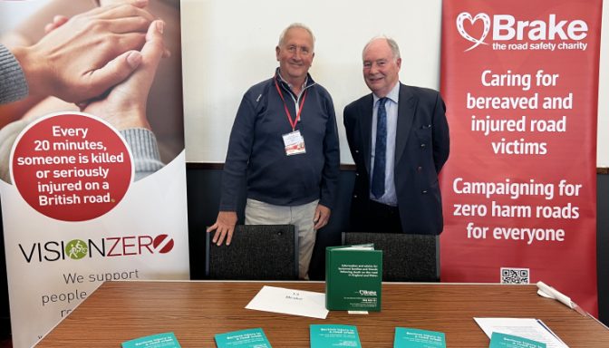 Two cheerful older men pose for a photo between two banners with Brake, the road safety charity's logo on.