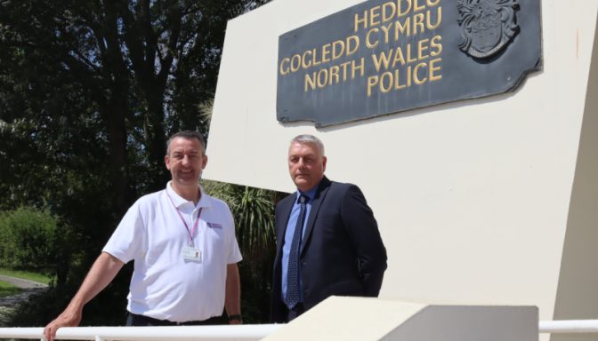 Two middle aged men stand outside a North Wales Police station, posing for a photo