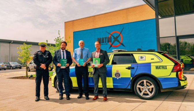 Three smartly-dressed men and one uniformed police officer posing for a photograph next to a police car. They are holding copies of a book with a green cover.