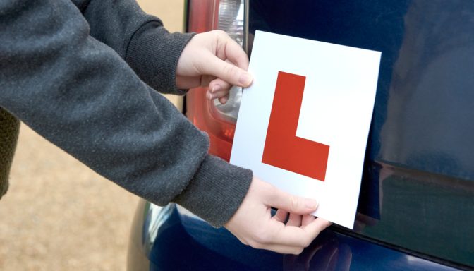 A young person attaching an L plate to the back of a car, alerting other road users that they are a learner driver.