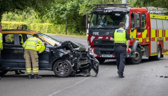 A road crash scene, with a damaged black car and a fire engine attending the scene. There is one police officer and one firefighter on the scene.