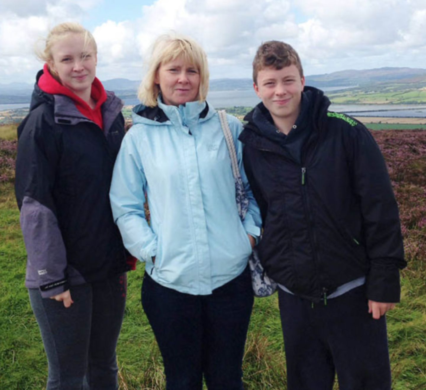 A teenage girl and boy pose for a photo with their mum. They are outdoors with a natural landscape visible behind them.