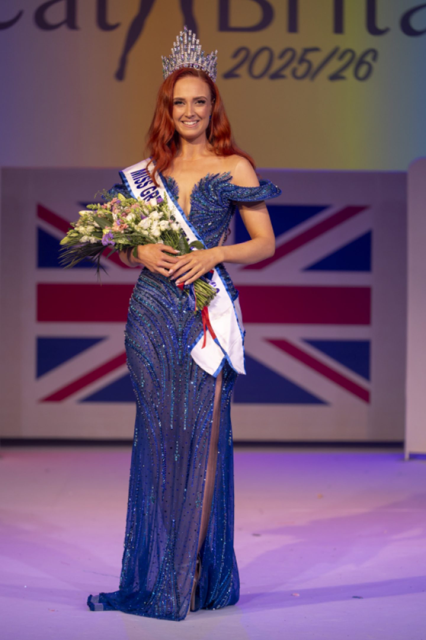 Alice poses for a photo after being crowned Miss Great Britain 2025/26. She is wearing a long, off-the-shoulder, sequined blue evening gown, a sash, and a crown, and holding a bouquet of flowers.