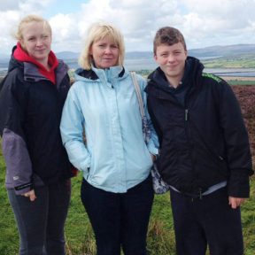 A teenage girl and boy pose for a photo with their mum. They are outdoors with a natural landscape visible behind them.