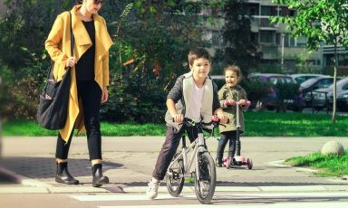 A woman and two children using a zebra crossing, The woman is walking, the bigger child is on a bike, and the smaller child on a scooter.