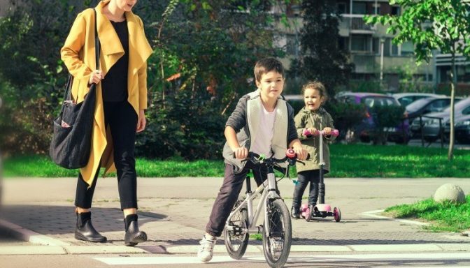 A woman and two children using a zebra crossing, The woman is walking, the bigger child is on a bike, and the smaller child on a scooter.