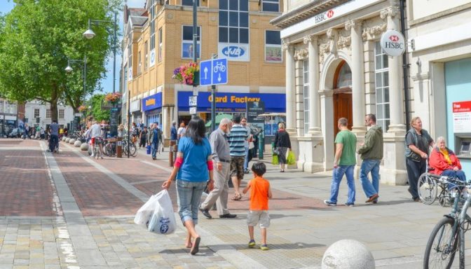 A street in a town. There is a blue bus parked at the bus stop on the left, and walking and cycle paths on the right. There are high street shops including a HSBC bank and a Card Factory. Many people are walking.