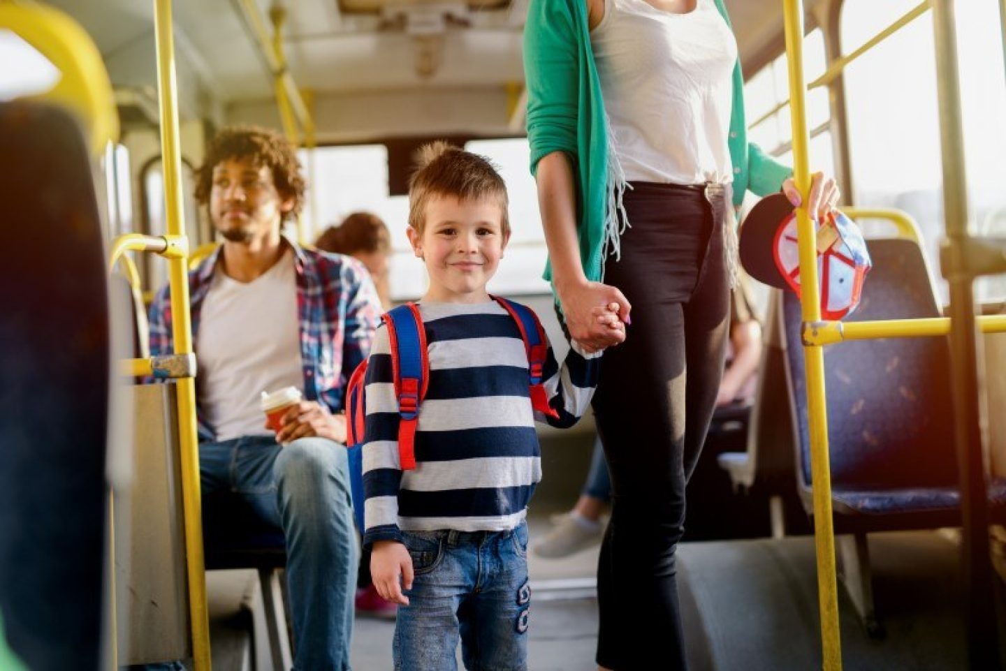 A young child is stood on a bus, holding his mother's hand. There are three other people behind them on the bus.