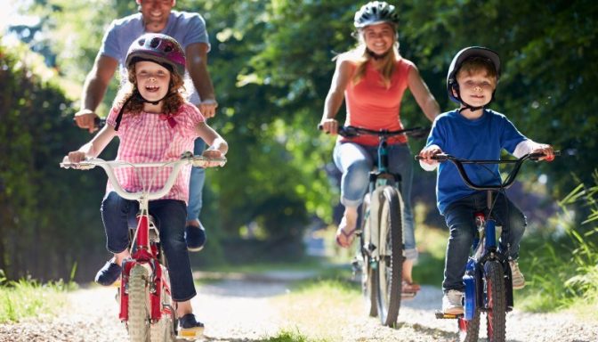 A man, woman, and two children are cycling. They are on a wide forest track, with no traffic, and are wearing helmets.