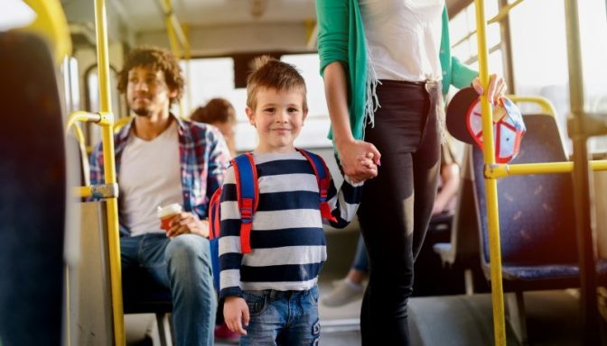 A young child is stood on a bus, holding his mother's hand. There are three other people behind them on the bus.