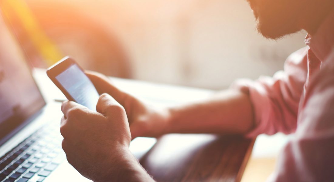 A person sat at a laptop, with their phone in their hands. They are looking at the phone screen.