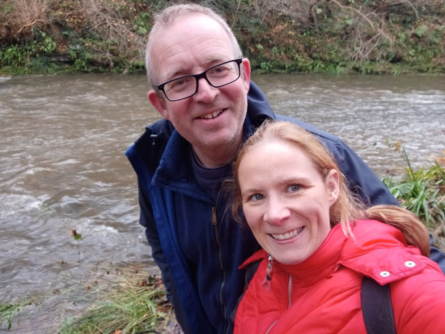 A man and woman pose for a selfie by a river. They're wearing outdoor gear so must be walking.
