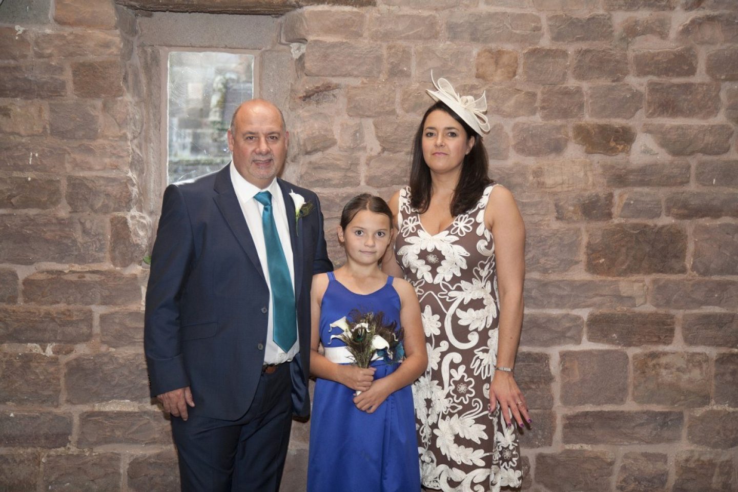 A mother and father pose for a photo with their young daughter. They are dressed formally, perhaps to attend a wedding,