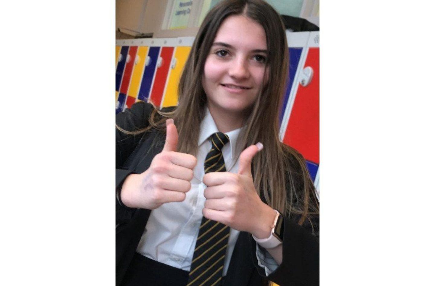 A young girl poses for a photograph in front of lockers in a school. She is smiling and holding both hands up in a 'thumbs up' gesture.