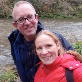 A man and woman pose for a selfie by a river. They're wearing outdoor gear so must be walking.