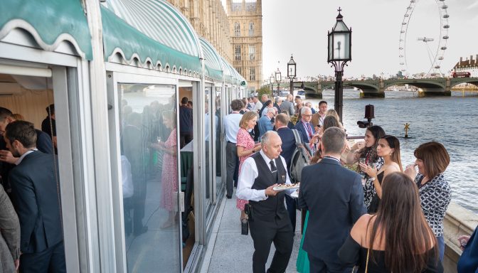 Smartly dressed people at an event at the Houses of Parliament. The photo is taken on a balcony, with people stood talking, eating and drinking. The River Thames is on the right, and the London Eye in the background